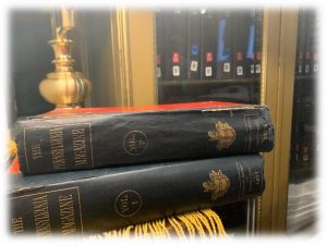 This is a photo of two leather bound volumes on top of each other with a light colored wooden bookshelf in the background 